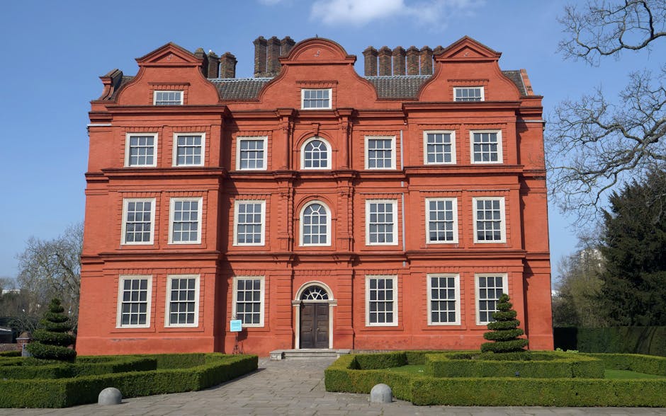 A large, multi-storey red brick building with a symmetrical façade featuring numerous white-framed sash windows arranged in three vertical columns. The building's central entrance has a dark wooden door topped with a semi-circular fanlight, and a small set of stone steps leads up to it. The roof has a distinctive design with multiple gables, chimney stacks, and decorative brickwork. In the foreground, there is a well-maintained garden with neatly trimmed box hedges forming rectangular patterns and two small, topiary trees on either side of the pathway. The garden is bordered by a paved walk, with large, rounded stone bollards placed along the edges. The surrounding environment includes a partly cloudy sky with patches of blue, and a few tall leafless trees are visible on the right side, suggesting a winter or early spring setting. The scene appears to be captured during daylight, with even lighting highlighting the building's architectural details and the lush greenery of the garden, which contrasts with the red brick façade, in an area that may be part of a historic estate or public park. The setting subtly aligns with themes of private estate management or on-site clearance, consistent with services like those offered by Waste Clearance Richmond.