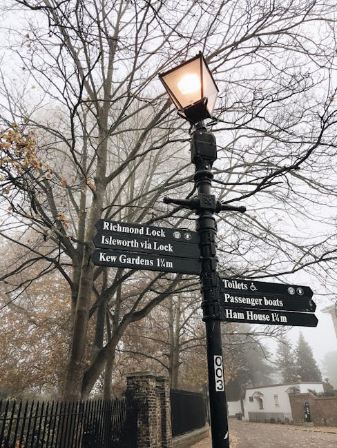 A black cast-iron lamppost standing on a paved sidewalk amidst leafless trees with bare branches. The lamppost features a traditional design with a glowing lantern at the top and multiple rectangular black directional signs attached to its pole, indicating local destinations such as Richmond Lock, Isleworth via Lock, Kew Gardens 1/4 mile, Toilets & Passenger Boats, and Ham House 1/4 mile. The surrounding environment includes a black metal fence on the left, a residential building and a low brick wall in the distance, with a foggy atmosphere reducing visibility of the background. The scene suggests an outdoor area in a historic or parkside setting, where independent or private waste clearance services, like those offered by Waste Clearance Richmond, might operate in proximity to public infrastructure, emphasizing the need for efficient rubbish removal and site clearance within such environments.