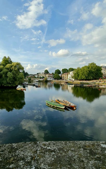 A serene riverside scene featuring a calm waterway reflecting a partly cloudy sky with patches of blue. Several small boats, varying in colour from green to beige, are docked or floating near the bank, with some appearing to be moored side by side and others slightly separated. The riverbank on the right has a mixture of mature trees with lush green foliage and residential buildings, including stone or brick houses, some with balconies or terraces overlooking the water. The background shows a gentle incline with more trees and additional buildings, creating a peaceful, semi-urban environment. The environment appears well-maintained, with no visible litter or debris, and the natural light suggests a bright day. This image, captured in a location where private or independent waste management might be relevant, exemplifies an outdoor setting where rubbish removal or waste clearance services could be employed to preserve the area's cleanliness and natural beauty, aligning subtly with alternative waste handling options in a community setting.